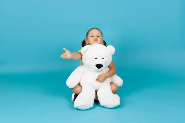 Een staartkind zit op de vloer gekruist, knuffelt een witte teddybeer en blaast een kus op een blauwe achtergrond — Stockfoto