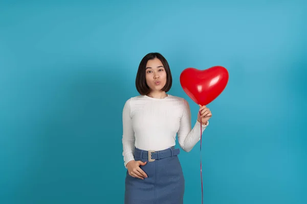 Portret van een jonge Aziatische vrouw blazen een kus en het houden van een rode hartvormige ballon geïsoleerd op een blauwe achtergrond — Stockfoto