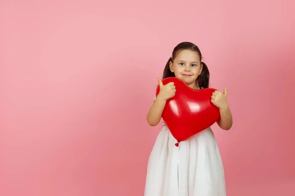 Burlarse amablemente, chica de corazón en vestido blanco sosteniendo globo rojo en forma de corazón, dando un pulgar hacia arriba en dos manos, aislado sobre un fondo rosa — Foto de Stock
