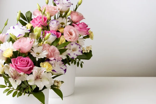 Farklı boyutlarda iki buket gül, papatya, lisianthus, kasımpatı, açılmamış tomurcuklar beyaz bir karton kutuda beyaz bir masa ve duvarda.