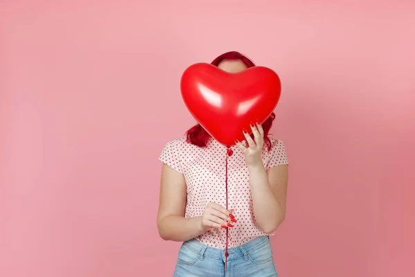Een vrouw met rood haar verbergt haar gezicht achter een grote vliegende rode hartvormige ballon in haar handen, geïsoleerd op een roze achtergrond — Stockfoto