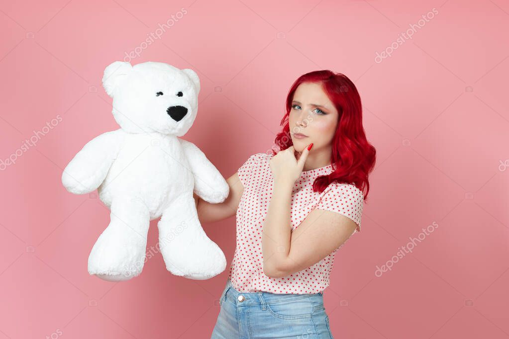 close-up brooding, suspicious young woman with red hair holds a large white teddy bear and rubs her chin with her hand , isolated on a pink background