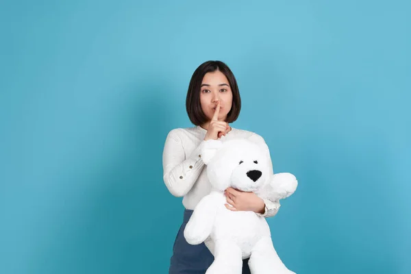 Vreugdevolle, geheimzinnige jonge aziatische vrouw houdt een grote witte teddybeer en een vinger op haar mond vragen om stilte, geïsoleerd op een blauwe achtergrond — Stockfoto
