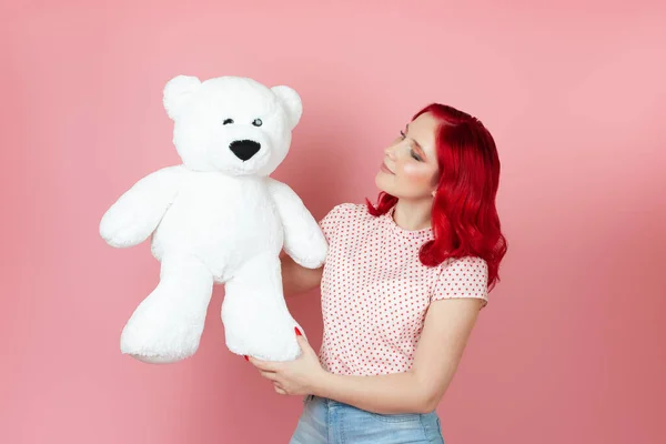 Close-up schattig in profiel jonge vrouw met rood haar kijkt naar een grote witte teddybeer geïsoleerd op een roze achtergrond — Stockfoto