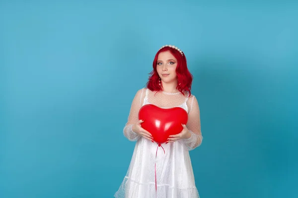 하얀 드레스를 입고 빨간 머리를 한 행복 한 젊은 여성이 양손으로 파란 배경에 심장 모양의 풍선을 들고 있다. — 스톡 사진