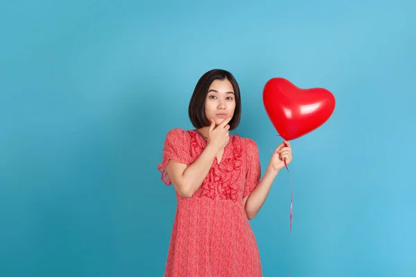 Mujer asiática joven y soñadora con un vestido rojo sostiene un globo rojo volador en forma de corazón y se frota la barbilla con la mano, aislada sobre un fondo azul — Foto de Stock