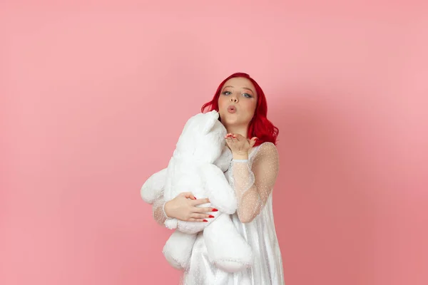 하얀 드레스를 입고 빨간 머리를 가진 귀엽고 귀여운 여성이 키스를 하고 핑크 색 배경에 고립 된 커다란 흰색 테디 베어를 껴안는다.. — 스톡 사진