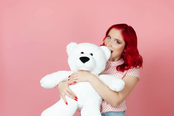 귀엽고 장난기많고 빨간 머리의 여자가 핑크 색 배경에 고립 된 커다란 흰색 곰 곰의 귀를 깨물었습니다. — 스톡 사진