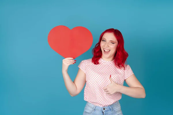 Retrato de una joven traviesa y juguetona con el pelo rojo sosteniendo un corazón de papel rojo, guiñando un ojo y dando un pulgar hacia arriba, aislado sobre un fondo azul — Foto de Stock