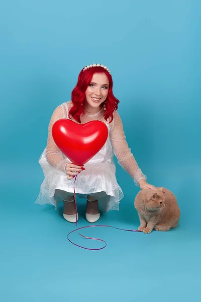 Una joven sonriente con un vestido blanco sostiene un globo volador en forma de corazón, se agacha y aprieta a un gato rojo británico, aislado sobre un fondo azul — Foto de Stock