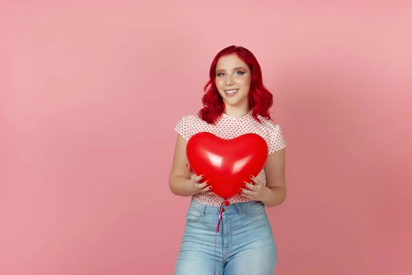 Primo piano sorridente, bella giovane donna con i capelli rossi tiene un grande palloncino rosso volante a forma di cuore isolato su uno sfondo rosa — Foto Stock