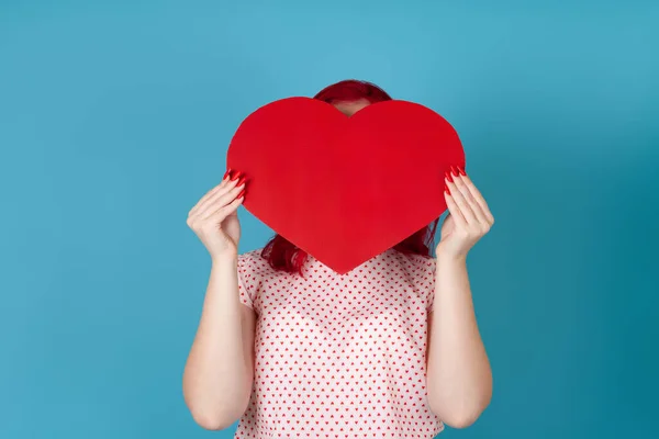 Mujer de pelo rojo oculta su cara detrás de un gran corazón de papel rojo aislado sobre un fondo azul — Foto de Stock