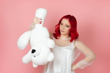 Üzgün, hayal kırıklığına uğramış, beyaz elbiseli, kızıl saçlı, büyük, beyaz bir oyuncak ayıyı pençesinden tutup pembe arka planda yalnız bırakan bir kadın.