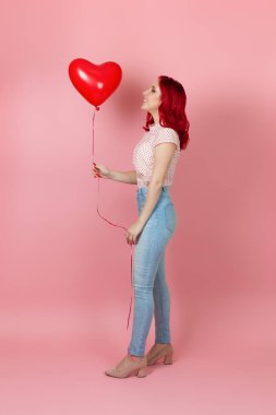 Uzun boylu, gülen, kızıl saçlı ve kot pantolonlu bir kadın elinde pembe bir arka planda izole edilmiş kırmızı bir balona bakıyor.