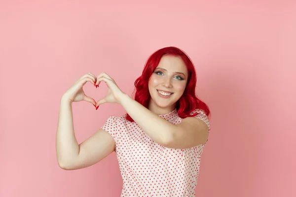 빨간 머리웃는 행복 한 여성이 핑크 색 배경에 고립 된 그녀의 손가락에서 심장 신호를 만들어 내고 있습니다. — 스톡 사진