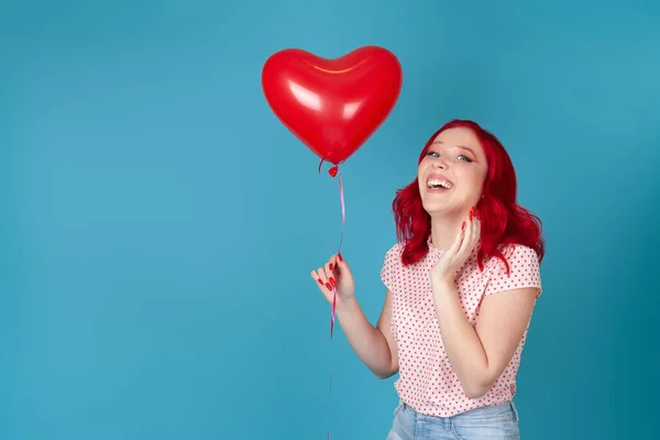 Retrato del estudio de una joven risueño regocijándose con el pelo rojo sosteniendo un globo volador rojo en forma de corazón, aislado sobre un fondo azul — Foto de Stock