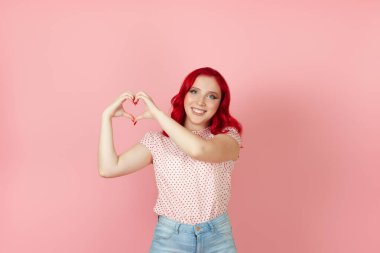Mutlu, gülen kızıl saçlı bir kadın pembe arka planda izole edilmiş parmaklarından kalp işareti yapar.