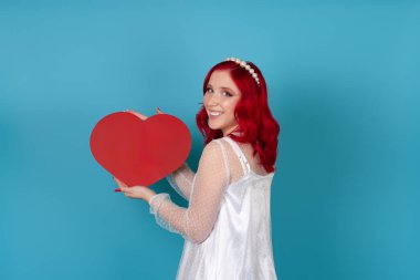 Neşeli, memnun bir kadın beyaz elbiseli, kızıl saçlı, mavi arka planda izole edilmiş kırmızı bir kalp taşıyor.