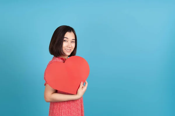 Feliz, sonriente joven asiática mujer en rojo vestido celebración grande rojo papel corazón como ventilador aislado en azul fondo — Foto de Stock