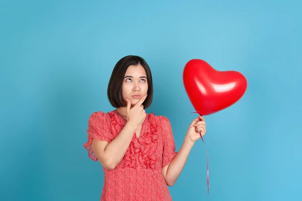 Una joven asiática dudosa y vacilante con un vestido rojo mira hacia los lados, sostiene un globo rojo volador en forma de corazón y se frota la barbilla con la mano, aislada sobre un fondo azul — Foto de Stock