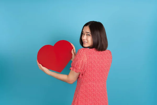 Glimlachen mooie jonge aziatische vrouw in rode jurk staat achteruit en houdt grote rode papieren hart geïsoleerd op blauwe achtergrond — Stockfoto