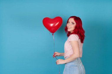 Yakın plan mutlu, dalgalı, kırmızı saçlı bir kadın elinde mavi arka planda izole edilmiş, kalp şeklinde kırmızı bir balon tutuyor.