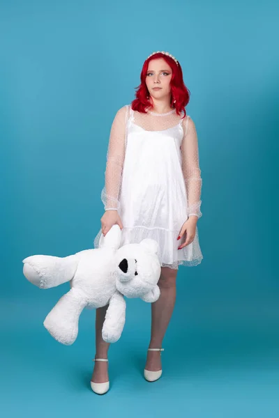 Mujer triste de cuerpo entero, decepcionado en un vestido blanco con el pelo rojo sostiene un oso de peluche blanco por la pata, aislado sobre un fondo azul — Foto de Stock