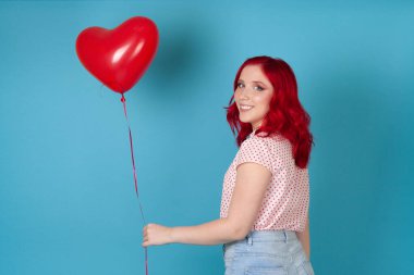 Kırmızı saçlı, gülümseyen güzel bir kadının portresi kalp şeklinde kırmızı bir balon tutuyor, mavi bir arka planda izole edilmiş.