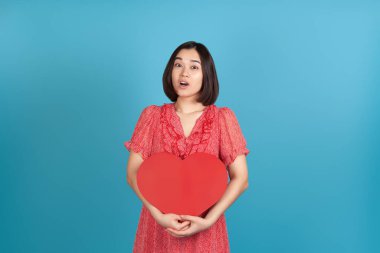 Şaşırmış, mutlu, heyecanlı, kırmızı elbiseli, ağzı açık Asyalı bir kadın. Elinde mavi arka planda izole edilmiş kırmızı bir kalp var.