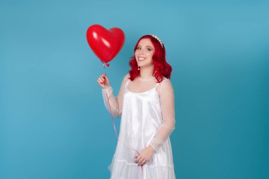 Kırmızı saçlı, beyaz elbiseli, güler yüzlü genç bir kadın elinde mavi arka planda izole edilmiş kalp şeklinde bir balon tutuyor.