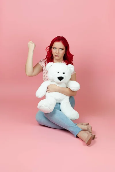 Обиженная, униженная женщина держит большого белого плюшевого медведя и трясет кулаком, изолированный на розовом фоне — стоковое фото