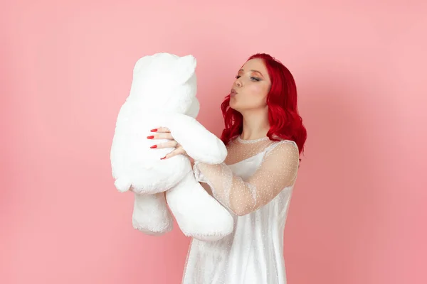 하얀 드레스를 입고 빨간 머리를 가진 귀엽고 귀여운 여성이 핑크 색 배경에 고립 된 커다란 흰색 테디 베어에게 키스를 건다. — 스톡 사진