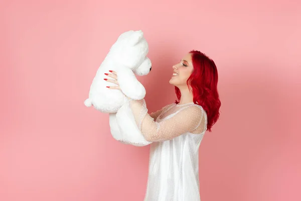 Portret in profiel van een vrolijke jonge vrouw in een witte jurk en met rood haar kijkend naar een grote witte teddybeer geïsoleerd op een roze achtergrond — Stockfoto