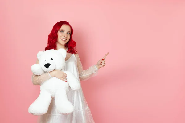 Een lachende, vrolijke jonge vrouw in een witte jurk en met rood haar houdt een grote witte teddybeer vast en wijst met haar wijsvinger naar een lege ruimte voor tekst, geïsoleerd op een roze achtergrond — Stockfoto