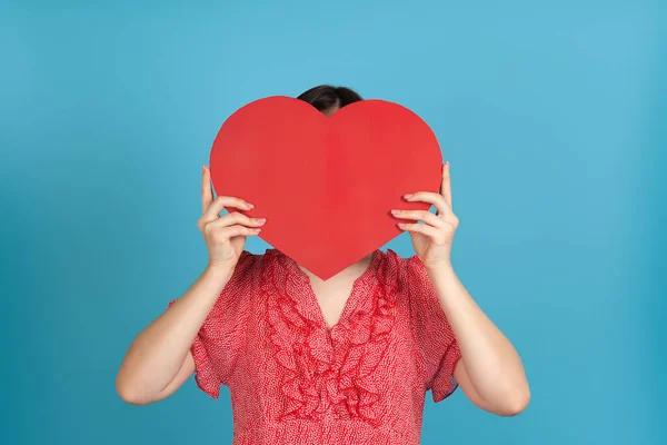 Mujer de pelo oscuro de cerca en un vestido rojo oculta su cara detrás de un gran corazón de papel rojo, aislado sobre un fondo azul — Foto de Stock