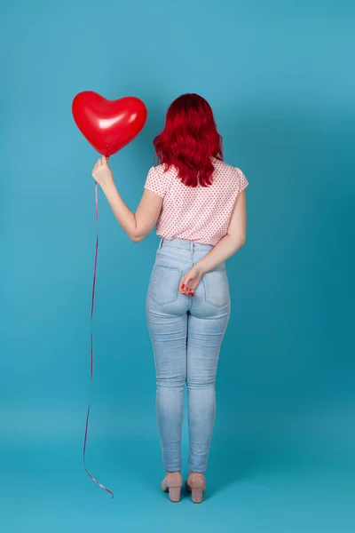 Pełnowymiarowa młoda kobieta w czerwonych włosach w dżinsach stoi plecami i trzyma w ręku czerwony latający balon w kształcie serca, odizolowany na niebieskim tle — Zdjęcie stockowe