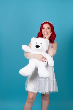 Şaşırtıcı genç bir kadın ağzı açık, beyaz elbiseli kırmızı dalgalı saçlı bir kadın büyük beyaz bir oyuncak ayıya sarılıyor, mavi bir arka planda soyutlanmış.