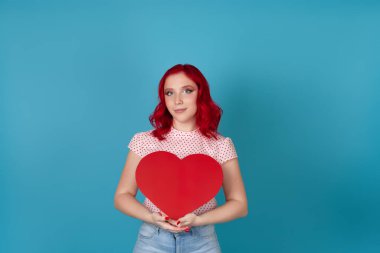 Sevimli, nazik, güzel, kızıl saçlı genç bir kadın iki elinde büyük kırmızı bir kalp tutuyor, mavi bir arka planda izole edilmiş.