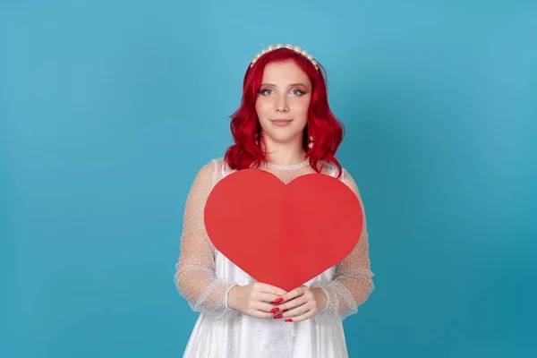 흰 드레스를 입고 빨간 머리에 빨간 종이 심장을 들고 파란 배경에 고립되어 있는 귀여운 젊은 여성 — 스톡 사진