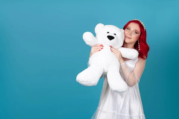 Close-up een mooie glimlachende vrouw in een witte jurk perst een grote witte teddybeer in haar gezicht, geïsoleerd op een blauwe achtergrond — Stockfoto