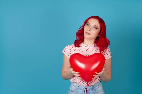 Retrato de una joven muy tierna con el pelo rojo sosteniendo un globo volador rojo en forma de corazón con ambas manos, aislado sobre un fondo azul — Foto de Stock