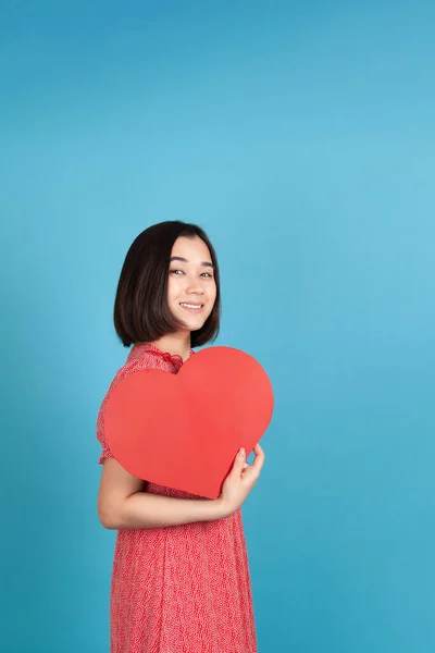 Alegre, alegre joven asiática mujer en rojo vestido celebración grande rojo papel corazón como ventilador aislado en azul fondo — Foto de Stock