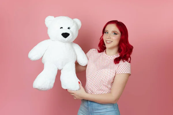 Linda, mujer romántica con el pelo rojo suavemente mimos un gran oso de peluche blanco aislado sobre un fondo rosa —  Fotos de Stock