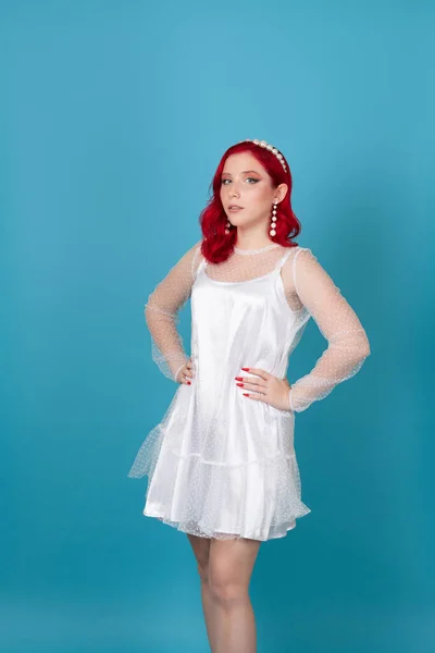 Delgada arrogante mujer joven segura de sí misma con el pelo rojo y las manos en la cintura en vestido de malla de seda blanca aislada sobre fondo azul — Foto de Stock