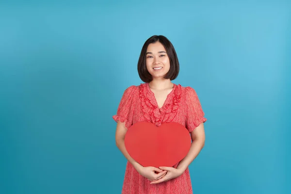 Mock-up een tevreden, charmante jonge Aziatische vrouw in een rode jurk houdt een groot rood papieren hart voor Valentijnsdag, geïsoleerd op een blauwe achtergrond — Stockfoto