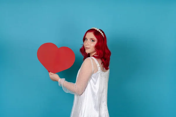 하얀 드레스를 입고 붉은 머리를 하고 파란 배경에 커다란 빨간 종이 심장을 들고 옆으로 서 있는 아름다운 여인 — 스톡 사진