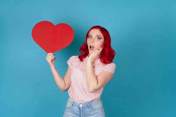 Una joven impresionada y sorprendida con el pelo rojo y la boca abierta sostiene un gran corazón de papel rojo aislado sobre un fondo azul — Foto de Stock