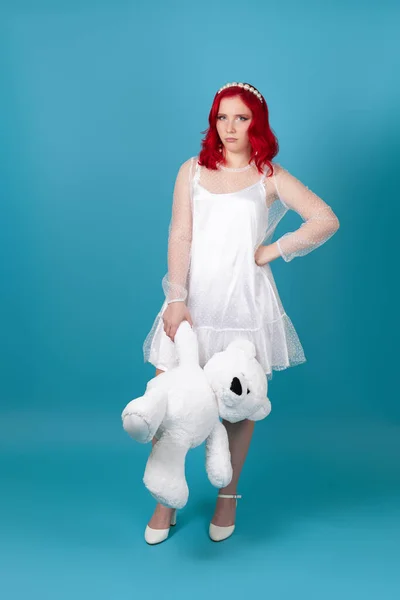 Mujer joven decepcionada, triste y resentida en vestido blanco con el pelo rojo sosteniendo un oso de peluche blanco por la pata, aislado sobre un fondo azul — Foto de Stock