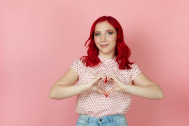 Yakın plan kızıl saçlı güzel bir kadın pembe arka planda izole edilmiş ellerinin parmaklarından kalp işareti yapıyor.