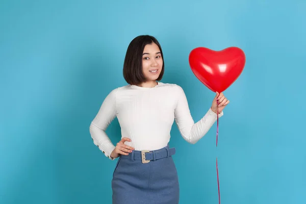 Flirten, ondeugende jonge Aziatische vrouw houdt een hartvormige vliegende ballon geïsoleerd op een blauwe achtergrond — Stockfoto